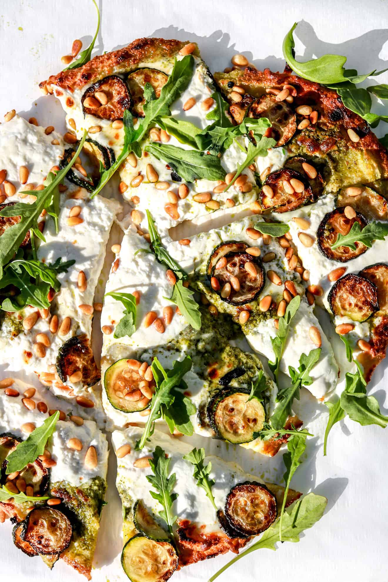 cauliflower flatbread topped with zucchini, burrata, and pine nuts cut into slices in sunlight on white background