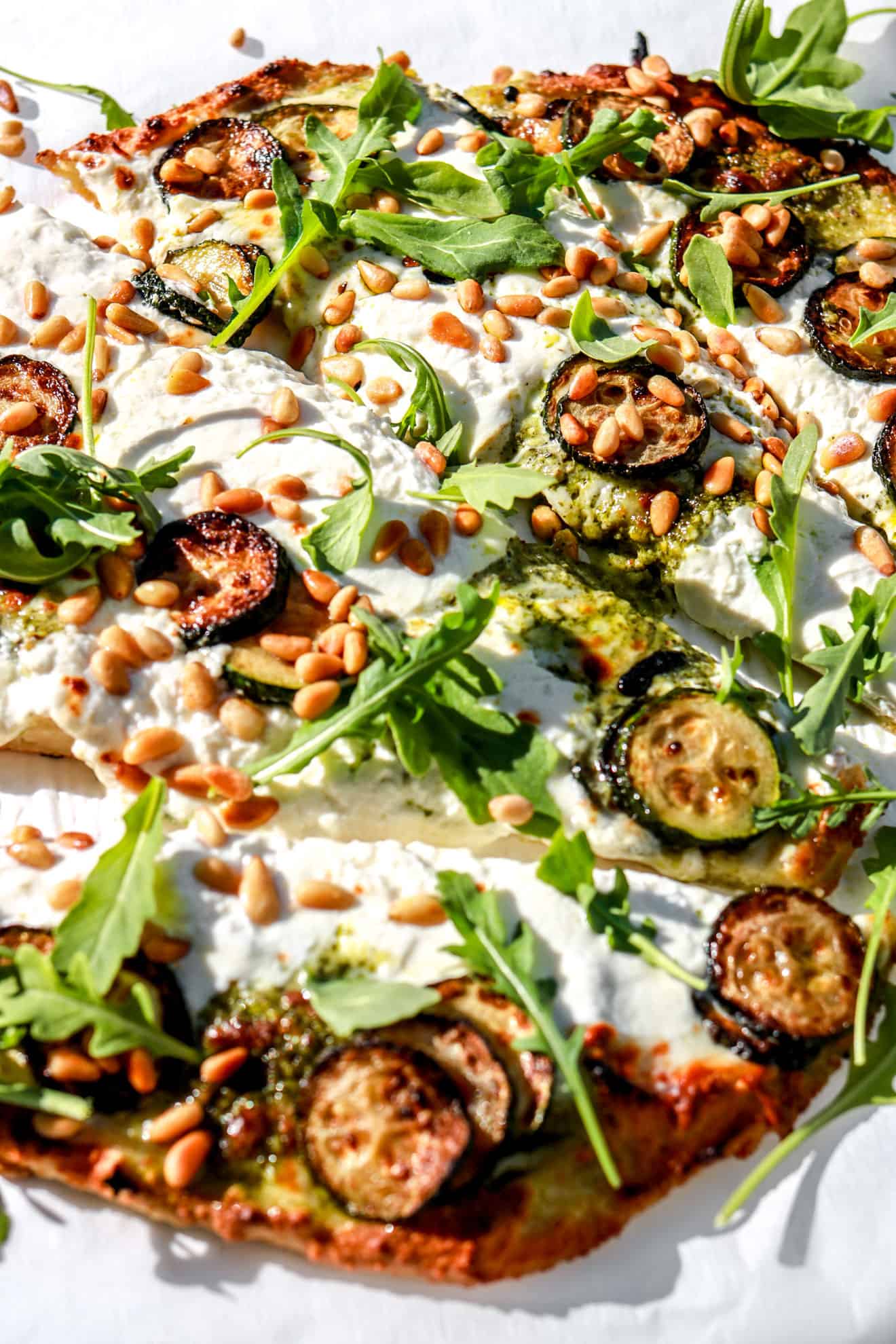 side view of cauliflower flatbread topped with zucchini, burrata, and pine nuts in sunlight on white background