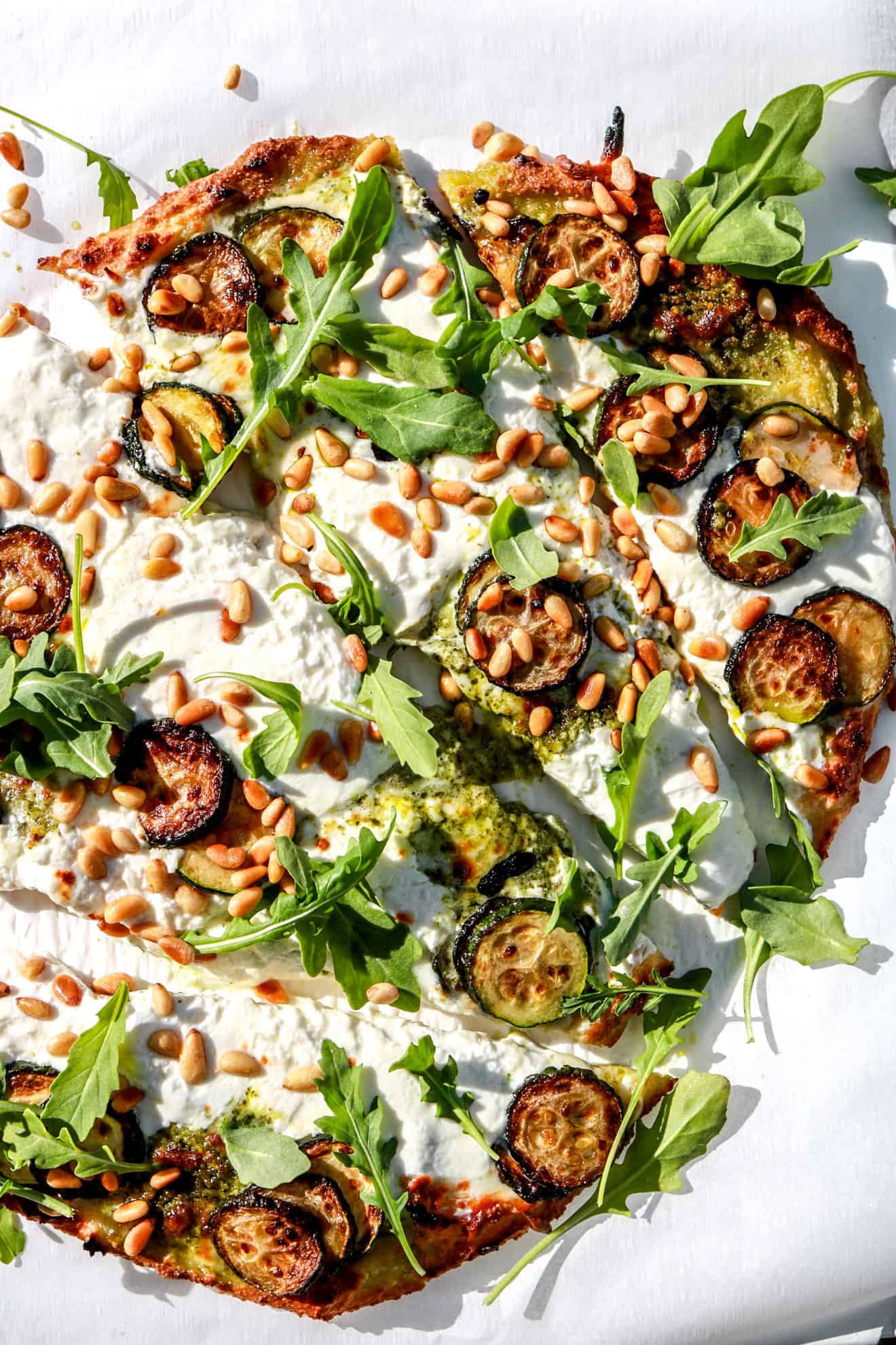 cauliflower low carb flatbread topped with zucchini, burrata, and pine nuts in sunlight on white background