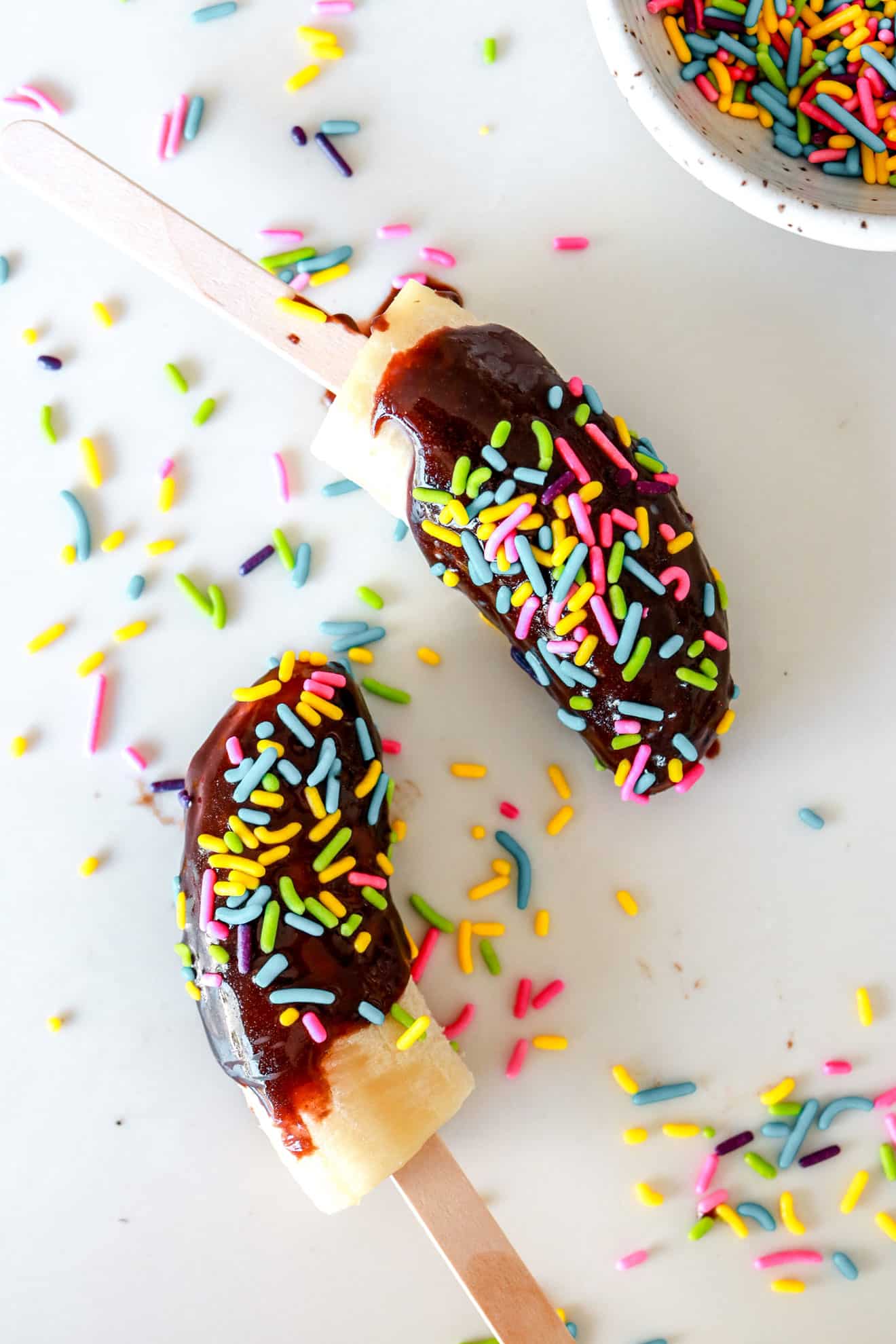 two Chocolate Covered Banana Pops on white counter with sprinkles