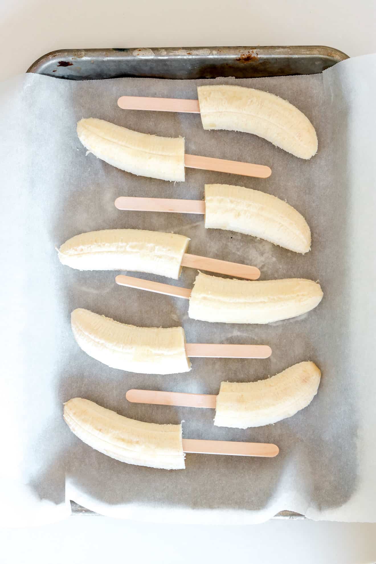 baking sheet lined with parchment paper and halved bananas with popsicle sticks process image for Chocolate Covered Banana Pops