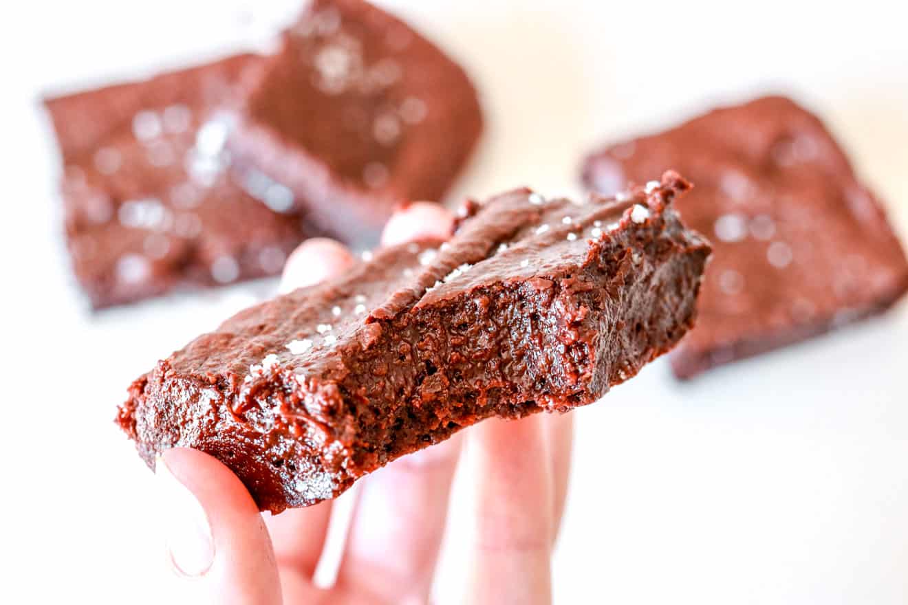 A hand is holding a fudgey almond flour brownie with a bite taken out of it. The brownie is sprinkled with flakey salt. More brownies are on the white counter, blurred in the background.