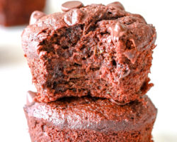 Two chocolate zucchini muffins are stacked on top of each other. The top muffin has a bite taken out of it. The muffins sit on a white surface with more chocolate muffins blurred in the background.