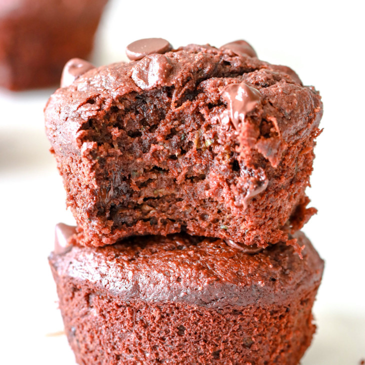 Two chocolate zucchini muffins are stacked on top of each other. The top muffin has a bite taken out of it. The muffins sit on a white surface with more chocolate muffins blurred in the background.