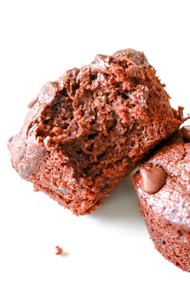 This is a side view of a chocolate muffin with a bite taken out. The muffin leans against another muffin. The muffins sit on a white counter. 