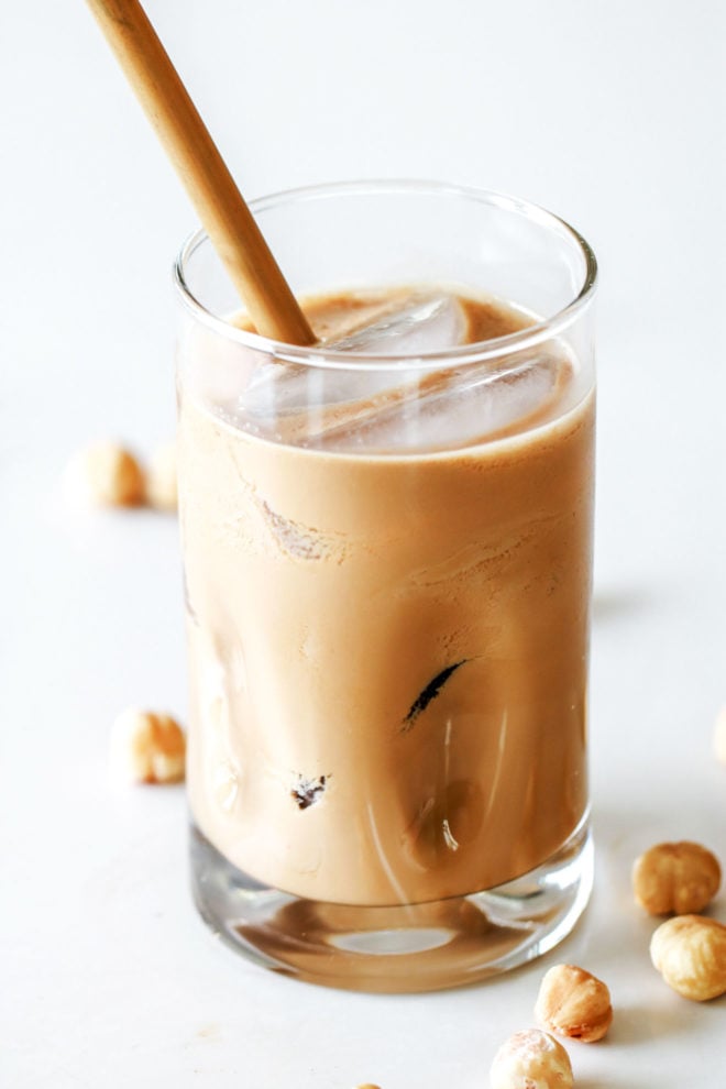 glass of iced hazelnut coffee with ice and a bamboo straw on a white counter with roasted hazelnuts around it