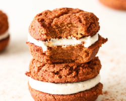 stack of two pumpkin whoopie pies, one with a bite taken out of it on a white counter with other pumpkin whoopie pies