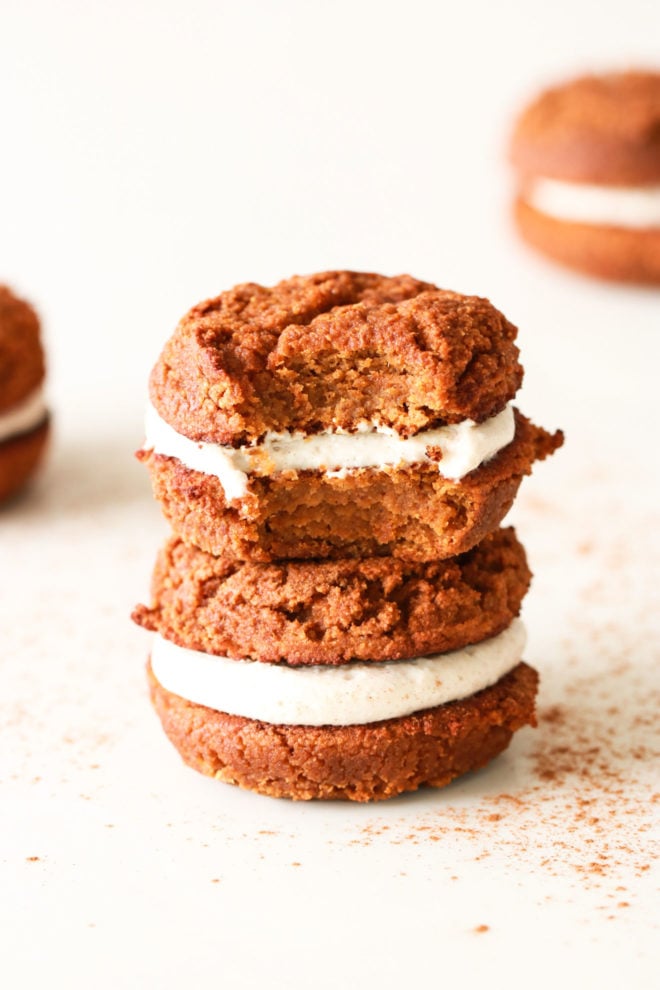 stack of two pumpkin whoopie pies, one with a bite taken out of it on a white counter with other pumpkin whoopie pies