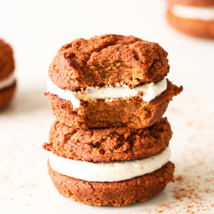 stack of two pumpkin whoopie pies, one with a bite taken out of it on a white counter with other pumpkin whoopie pies