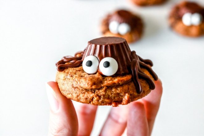 hand holding peanut butter cookie with an upside down peanut butter cup, candy eyeballs and chocolate legs to appear like a spider