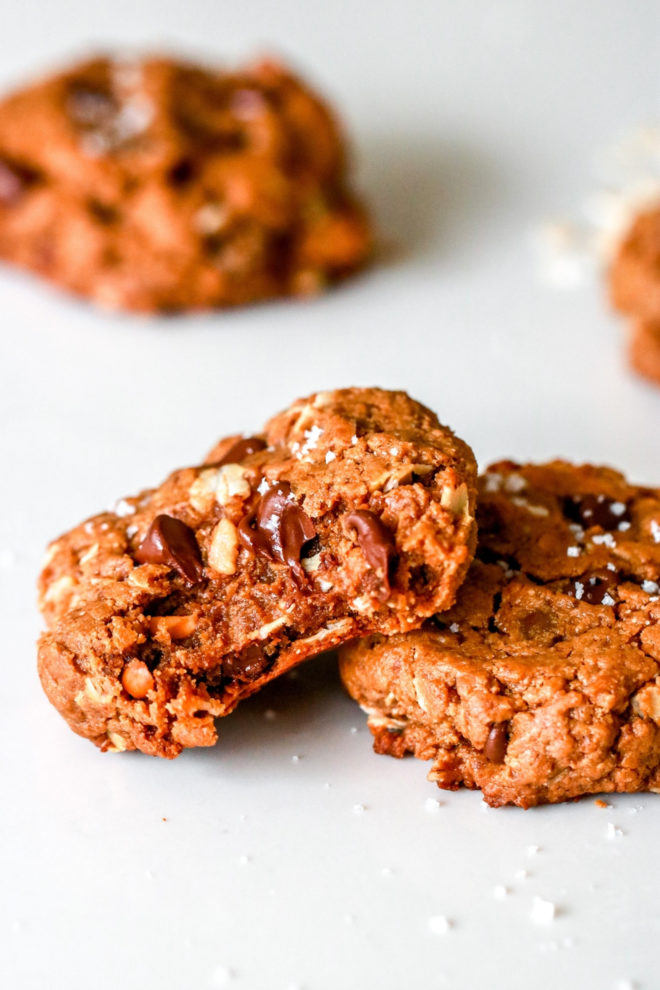 side view of a chocolate chip peanut butter oatmeal cookie with chocolate chips, chopped nuts, and oats with a bite taken out, leaning on its side against another cookie on a white counter with other cookies blurred in the background