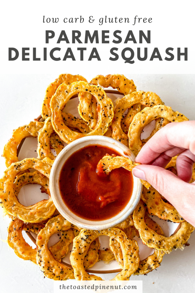 overhead image of a plate with delicata squash crusted with paremsan and herbs. a small bowl of marinara sauce is in the middle of the plate and a hand is dipping one of the squash rings into the marinara sauce. 