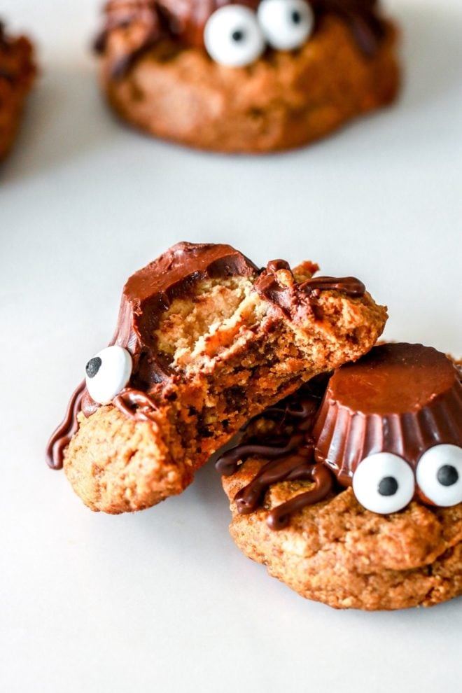 two peanut butter cookies decorated like spiders with a peanut butter cup and candy eyeballs, one with a bite taken out