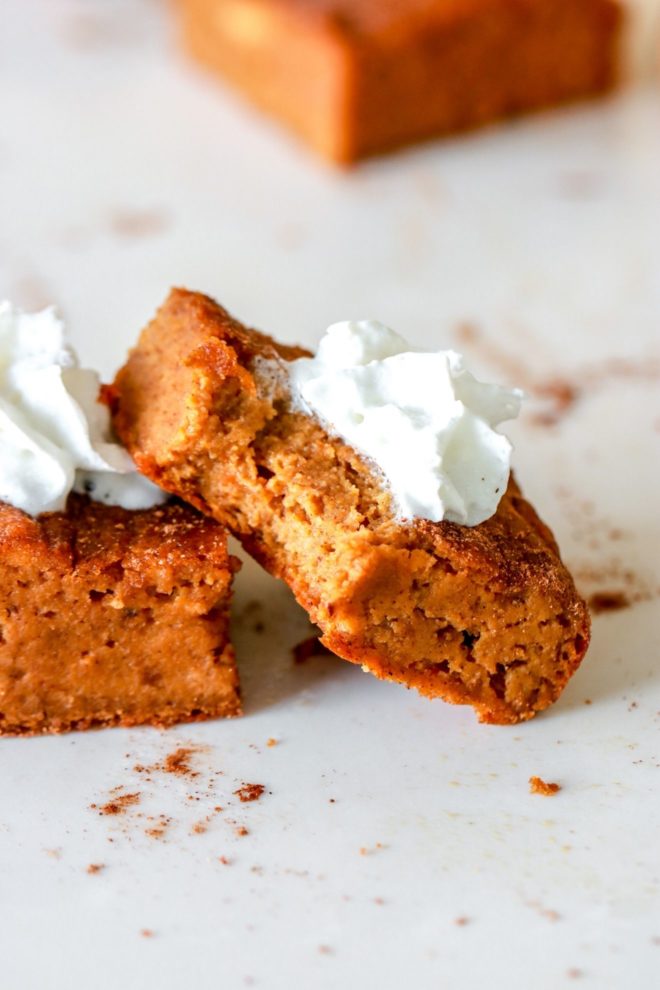 side view of pumpkin banana cake bar with a bite taken out leaning against another pumpkin banana cake bar on white counter with dollop of whipped cream