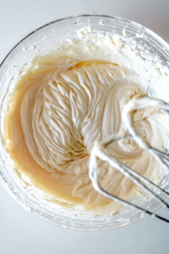 glass bowl with cream cheese frosting being whipped by an electric mixer