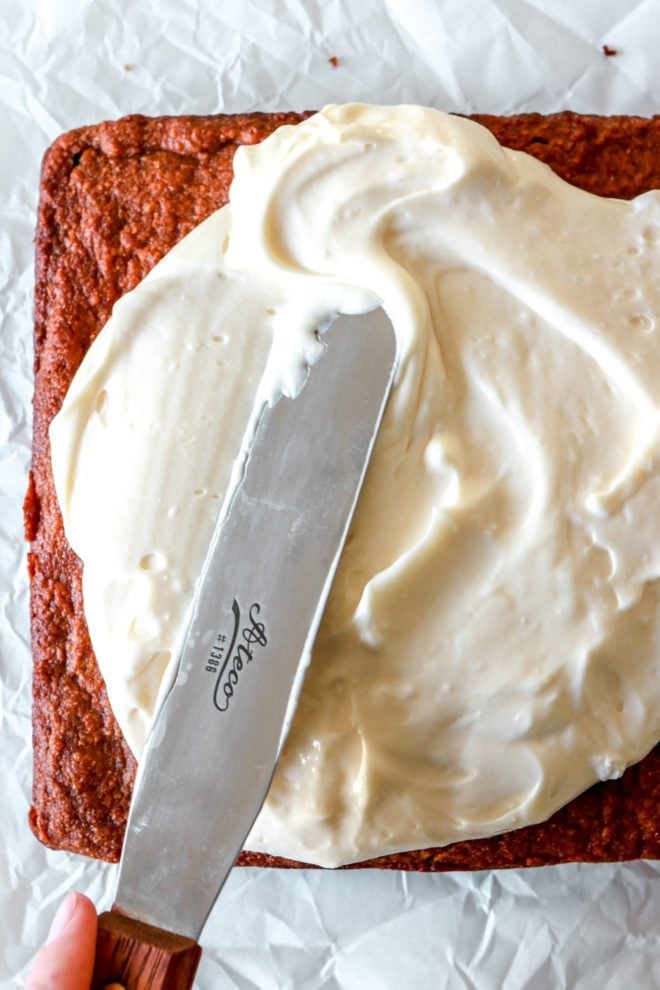 Spatula spreading out cream cheese frosting on top of a gluten free pumpkin cake on white parchment paper