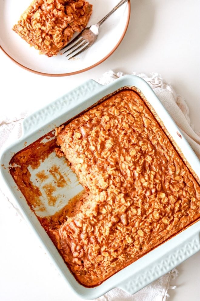 overhead image of a light blue dish with baked pumpkin oatmeal on a white counter with a slice taken out and placed on a white plate next to it