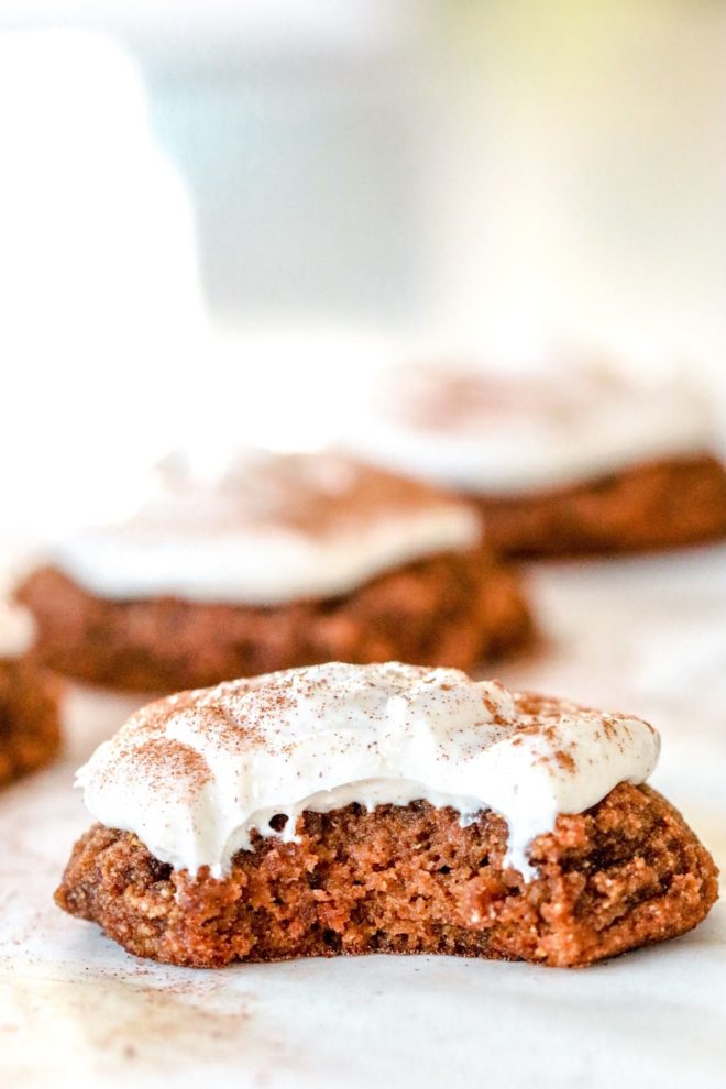 side view of a pumpkin cookie with cream cheese icing and a cinnamon sprinkle with a bite taken out and sitting on a white counter