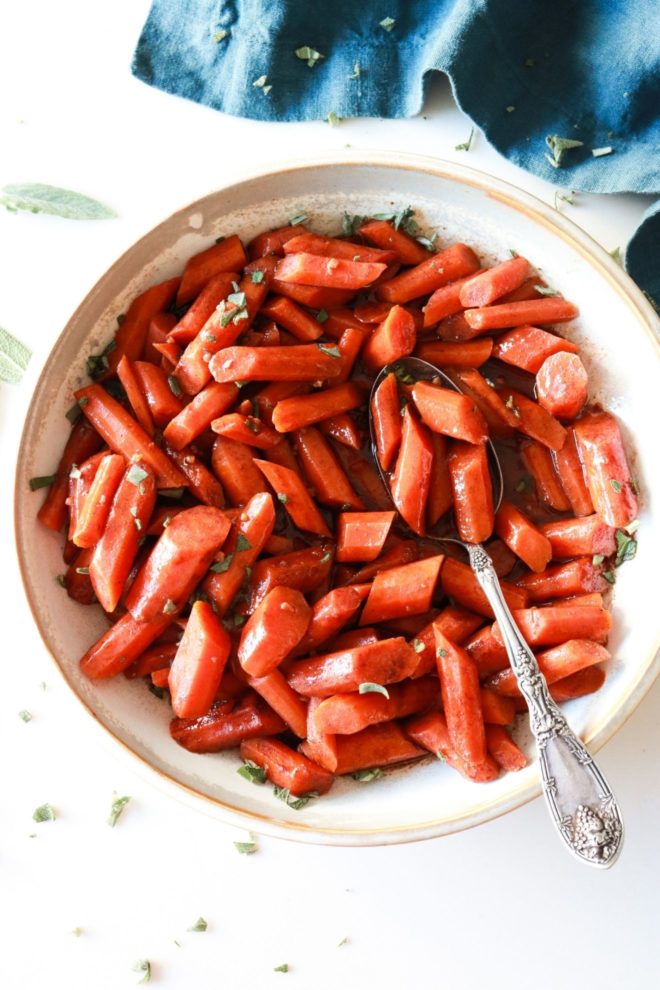 bowl with sweet cinnamon slow cooker carrots topped with chopped sage and a serving spoon laying inside the bowl