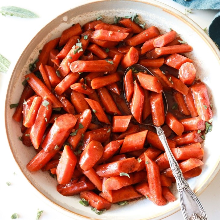 bowl with sweet cinnamon slow cooker carrots topped with chopped sage and a serving spoon laying inside the bowl