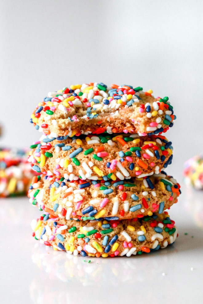 side view of a stack of four sprinkle cookies in a white counter, the top cookie has a bite taken out