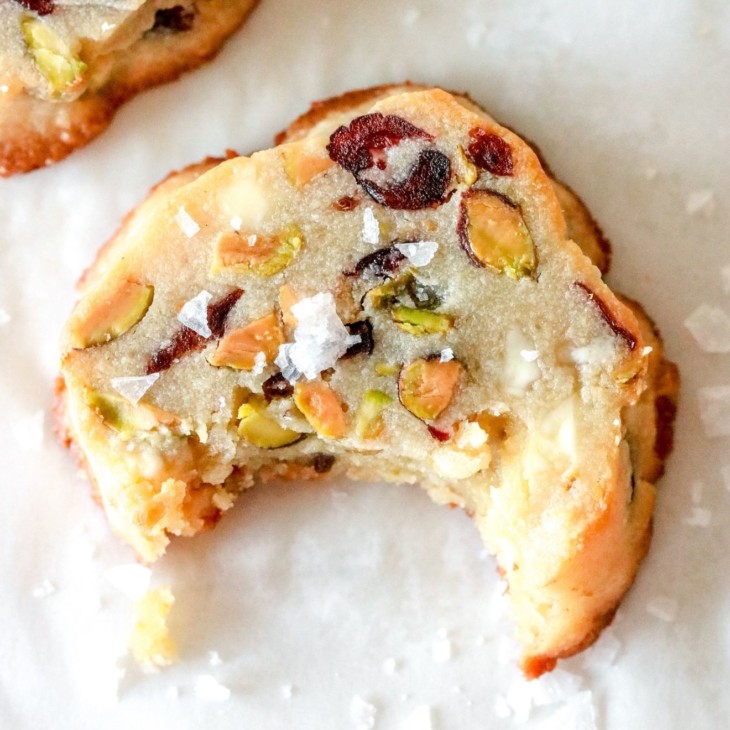 overhead image of shortbread cookie with cranberries, pistachios, and white chocolate pieces in it and salt sprinkled on top. a bite is taken out of the cookie and the cookie sits on a white counter.