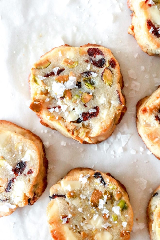overhead image of shortbread cookies with pieces of cranberry, pistachio, and white chocolate in the. The cookies are sprinkled with flaked salt and sit on white parchment paper.