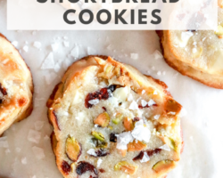 overhead image of shortbread cookie with cranberries, pistachios, and white chocolate pieces in it and salt sprinkled on top. a bite is taken out of the cookie and the cookie sits on a white counter. text overlay "low carb & gluten free white chocolate cranberry pistachio shortbread cookies"