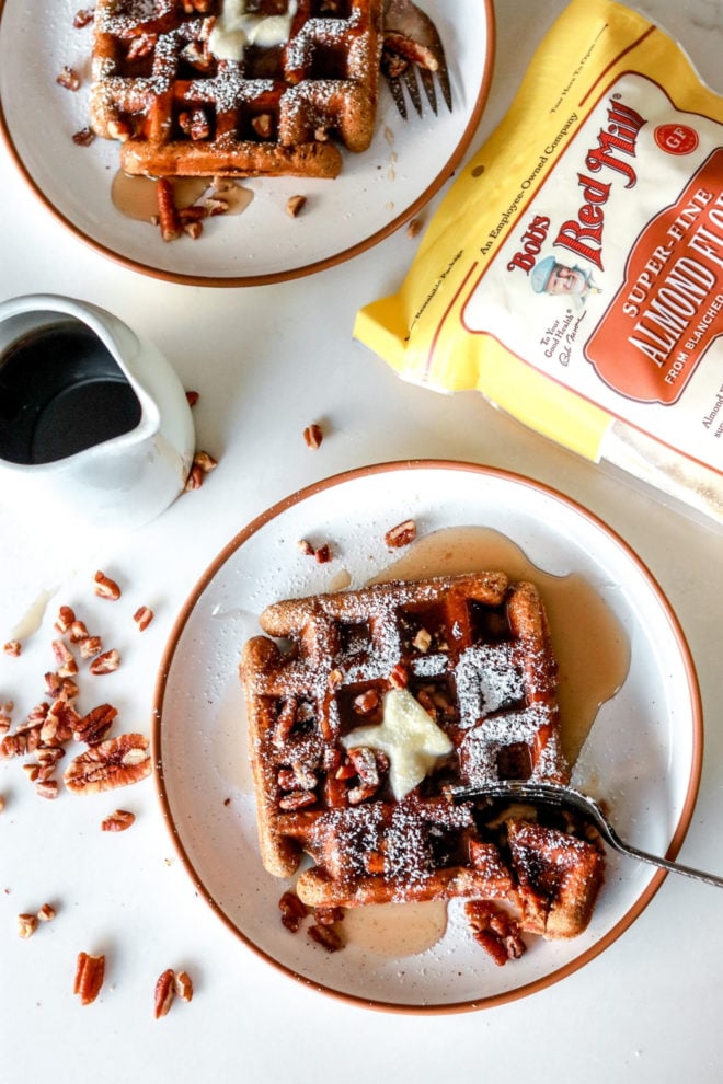 overhead image of white plates with gluten free gingerbread waffles topped with butter, syrup, powdered sugar, and pecans. A bag of bob's red mill almond flour lays on the counter next to the plates.