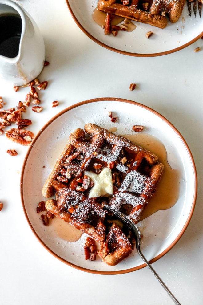 overhead image of white plates with gluten free gingerbread waffles on them. waffles are topped with butter, syrup, pecans, and powdered sugar.