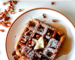 overhead image of a waffle on a white plate on a white counter. the waffle has butter, syrup, chopped pecans, and powdered sugar on top. A fork is cutting off a corner of the waffle.