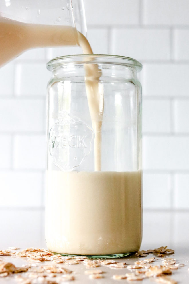 Side view of a glass pitcher pouring oat milk into a glass weck jar. jar sits on a white counter with oats scattered around it and white subway tile in the background.