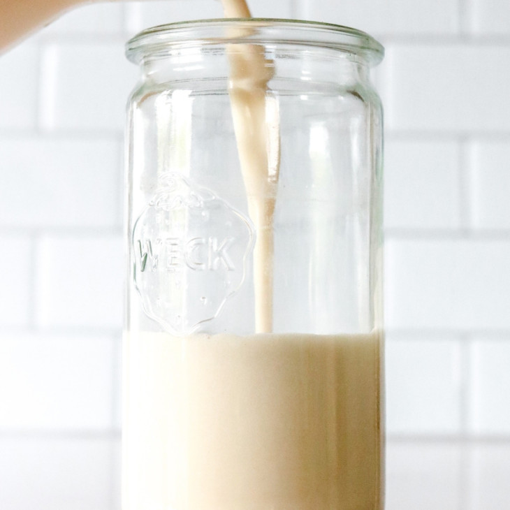 Side view of a glass pitcher pouring oat milk into a glass weck jar. jar sits on a white counter with oats scattered around it and white subway tile in the background.