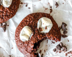 overhead image of a chocolate cookie with marshmallows on top and a bite taken out. cookie is on a white parchment paper and chopped chocolate scattered around the cookie.