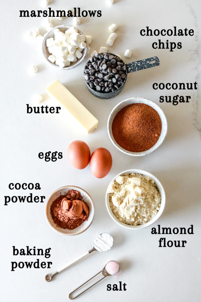 overhead image of ingredients on a white counter: marshmallows, chocolate chips, butter, coconut sugar, eggs, almond flour, cocoa powder, baking powder, and salt