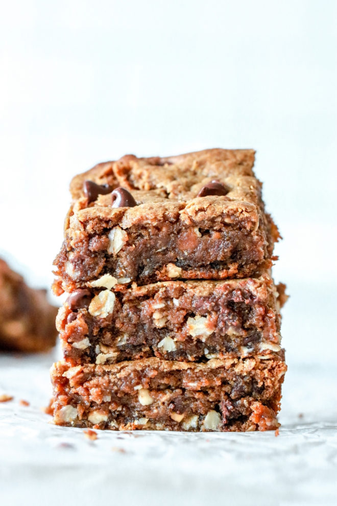 stack of three peanut butter bars loaded with chocolate chips, oats, and peanuts. The stack sits on a white counter and a white background, crumbs and other cookie bars are blurred in the background.