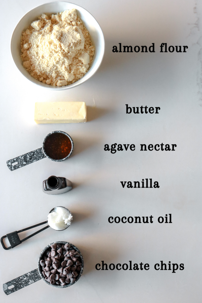 overhead image of almond flour, butter, agave nectar, vanilla, coconut oil, and chocolate chips. ingredients to make almond flour shortbread cookies are laid out on a white counter.