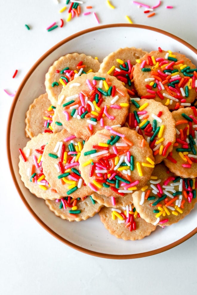 overhead image of a plate on a white counter. the plate has shortbread sprinkle cookies on it. rainbow sprinkles are scattered on the counter.