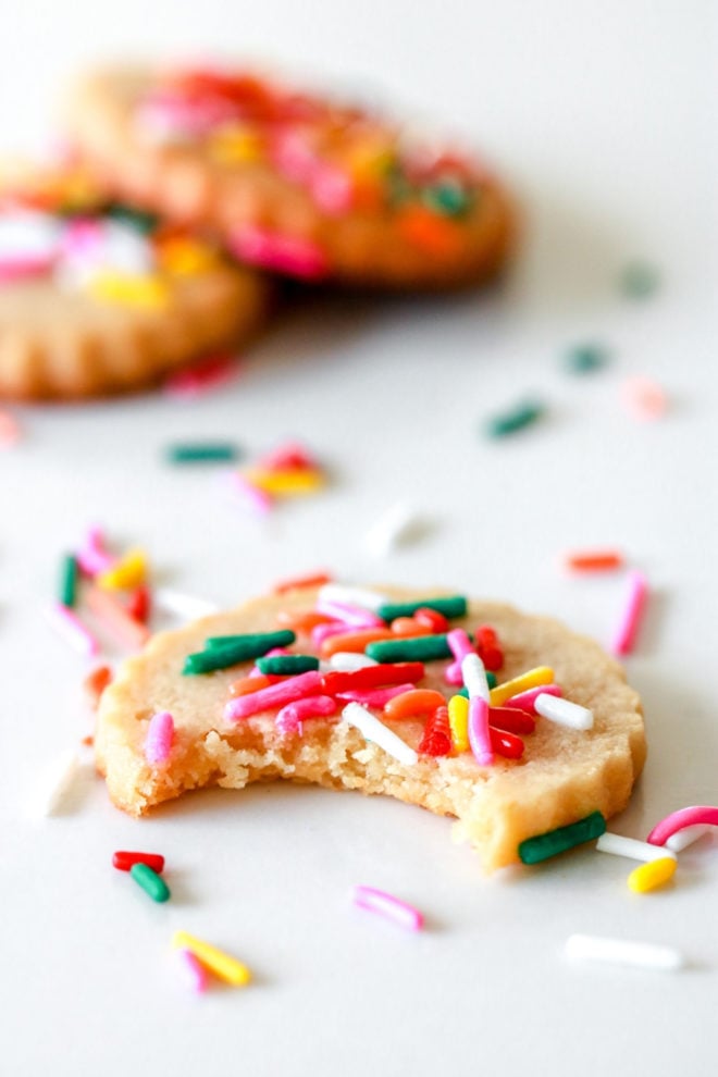 side view of a shortbread sprinkle cookie with a bite taken out of it. the cookie sits on a white counter with rainbow sprinkles scattered around it and two other sprinkle cookies blurred in the background.