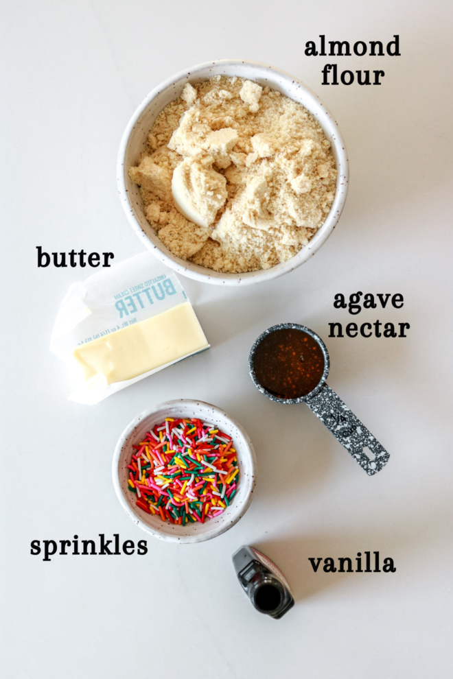 overhead image of almond flour, butter, agave nectar, sprinkles, and vanilla extract on a white counter