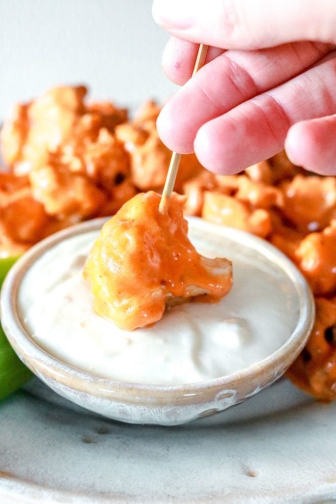 A hand holds a toothpick that has a cauliflower floret coated in buffalo sauce and dipped in a creamy blue cheese dressing. A plate of more buffalo cauliflower is blurred in the background.