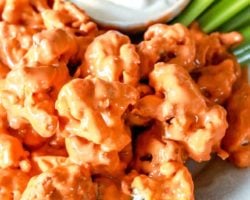 This is a side view and close up image of cauliflower florets on a plate. The florets are coated in buffalo sauce. A small bowl of creamy dressing and celery is in the background.