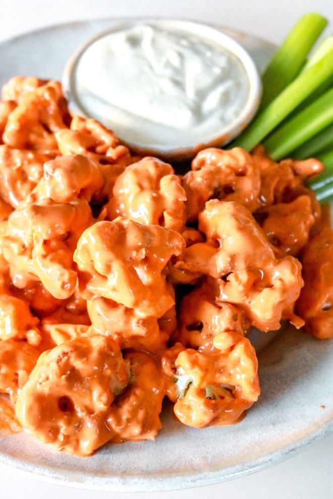 This is a side view and close up image of cauliflower florets on a plate. The florets are coated in buffalo sauce. A small bowl of creamy dressing and celery is in the background.