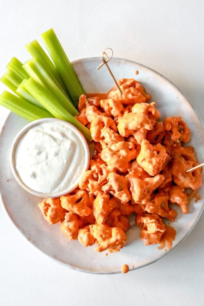 This is an overhead image of a plate with buffalo cauliflower florets. The plate also has a small bowl of blue cheese dip and celery sticks. Toothpicks are stuck into the florets.