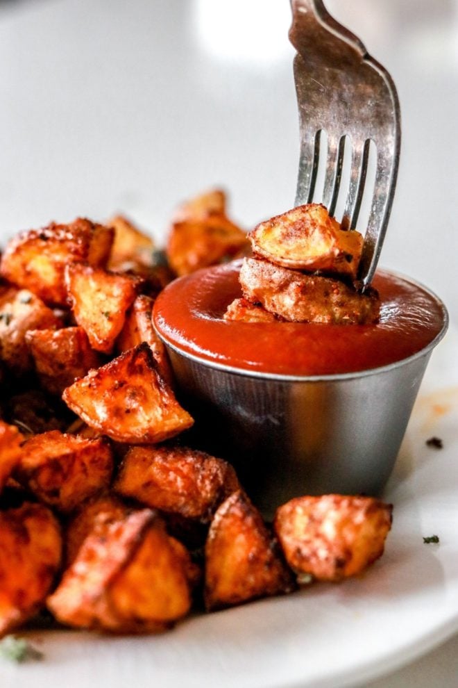 This is a side image of a fork dipping potatoes into a small bowl of ketchup. The ketchup bowl sits on a white plate along with other roasted potatoes.