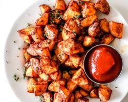 This is an overhead image of a white plate filled with roasted potatoes and a small bowl of ketchup.