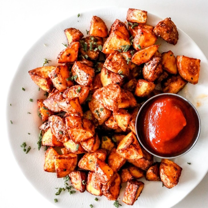 This is an overhead image of a white plate filled with roasted potatoes and a small bowl of ketchup.