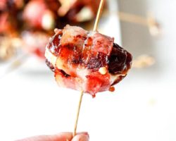 This image shows a hand holding a toothpick that has a bacon wrapped date on it. The date is stuffed with cheese and a plate of more dates is blurred in the background.
