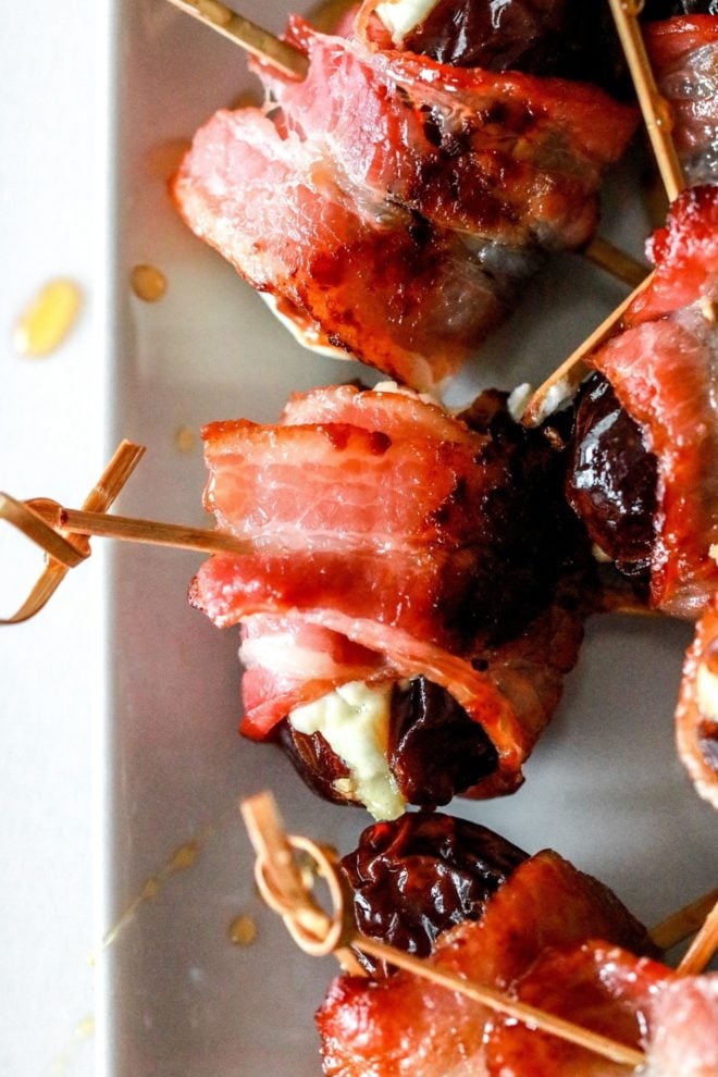 This is an overhead closeup image of bacon wrapped dates stuffed with cheese. The dates are skewered with toothpicks and lay on a white plate.