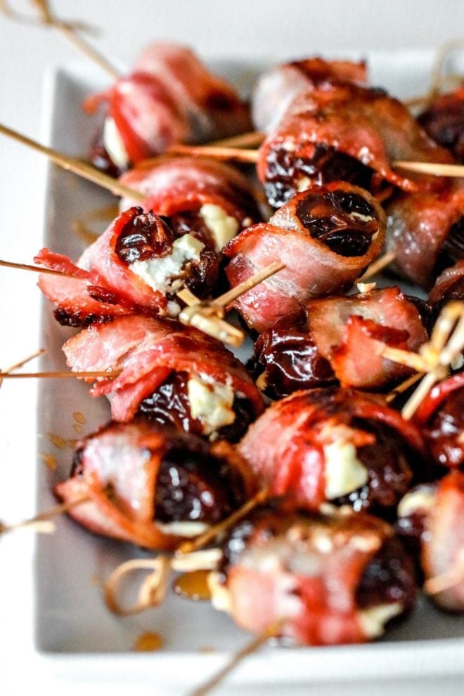 This is a side view of a white rectangle plate with a bunch of bacon wrapped dates on it. The dates are skewered with toothpicks and the plate sits on a white counter.